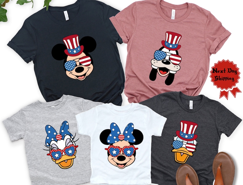 May include: Set of five t-shirts featuring cartoon characters wearing patriotic themed hats and sunglasses. The characters include Mickey Mouse, Minnie Mouse, Donald Duck, and Goofy. The shirts are in various colors, including navy blue, gray, white, and pink.