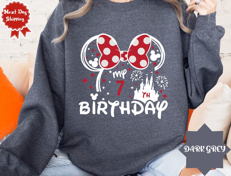 May include: Dark grey sweatshirt with a white graphic of Minnie Mouse ears, a red bow with white polka dots, a castle, fireworks, and the text "My 7th Birthday".