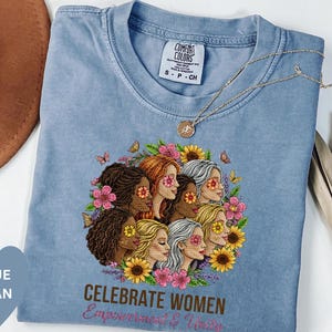 May include: A blue jean Comfort Colors t-shirt featuring a graphic of diverse women surrounded by flowers and butterflies. The text "CELEBRATE WOMEN Empowerment & Unity" is printed below the graphic. A gold necklace is draped across the shirt.
