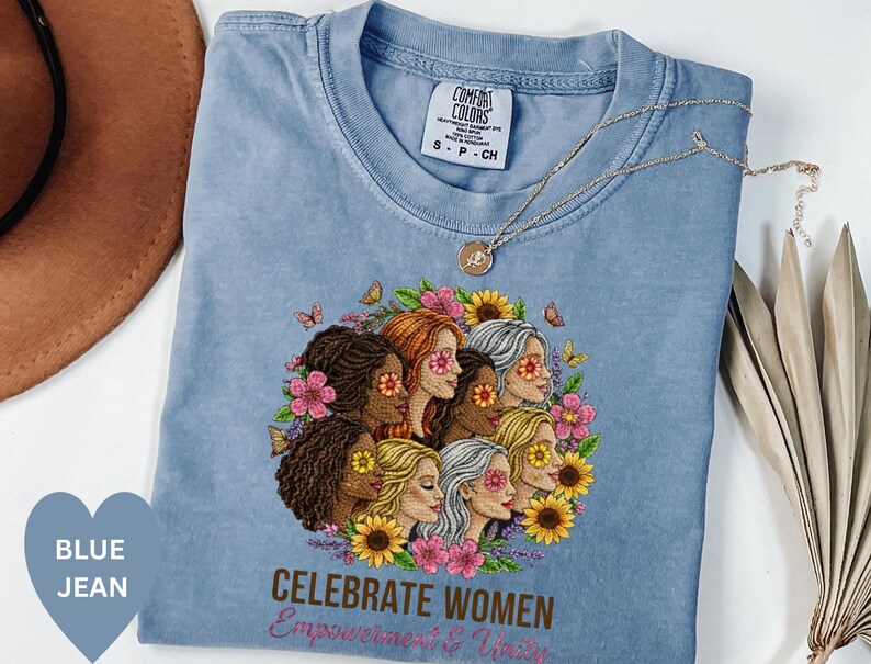 May include: A blue jean Comfort Colors t-shirt featuring a graphic of diverse women surrounded by flowers and butterflies. The text "CELEBRATE WOMEN Empowerment & Unity" is printed below the graphic. A gold necklace is draped across the shirt.
