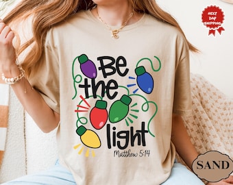 Peut inclure: T-shirt couleur sable avec l'inscription "Be the light" et le verset biblique Matthieu 5:14. Le motif comprend des guirlandes lumineuses de Noël de couleur violette, bleue, rouge, verte et jaune.