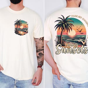 Pu&ograve; includere: T-shirt color crema con una scena di spiaggia al tramonto con una palma, onde e uccelli. La parte posteriore della maglietta mostra il testo "Forever Chasing Sunsets" e "Chasing Sunsets" in un font stilizzato, con la stessa grafica della spiaggia.
