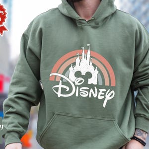 Puede incluir: Una sudadera con capucha de color verde militar que presenta un gr&aacute;fico blanco de la silueta de un castillo sobre un arco&iacute;ris de tonos apagados. La palabra Disney est&aacute; impresa en una fuente cursiva blanca debajo del castillo. Bolsillo frontal.