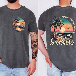 Pu&ograve; includere: T-shirt grigio scuro con grafica anteriore e posteriore. La grafica anteriore &egrave; un disegno circolare con una palma, un tramonto e una scena oceanica. La grafica posteriore dice "Forever Chasing Sunsets" e "Chasing Sunsets" con la stessa scena.
