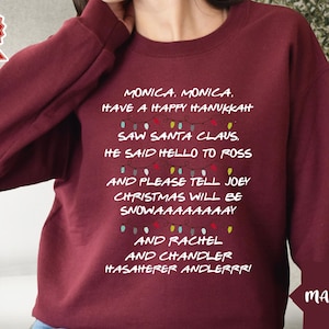 Puede incluir: Sudadera color granate con un estampado gráfico blanco de una cadena de luces de Navidad con el texto "MONICA, MONICA. HAVE A HAPPY HANUKKAH... SAW SANTA CLAUS. HE SAID HELLO TO ROSS AND PLEASE TELL JOEY CHRISTMAS WILL BE SNOWAAAAAAAAY... AND RACHEL AND CHANDLER HASAHERER ANDLERRRI"