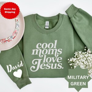 May include: Military green sweatshirt with the text "cool moms love Jesus" in white. The sleeve has the name "David" and a white heart. The sweatshirt is a crew neck style.