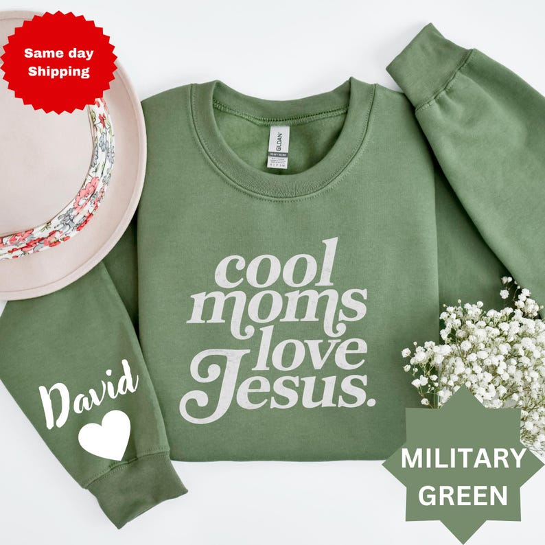 May include: Military green sweatshirt with the text "cool moms love Jesus" in white. The sleeve has the name "David" and a white heart. The sweatshirt is a crew neck style.