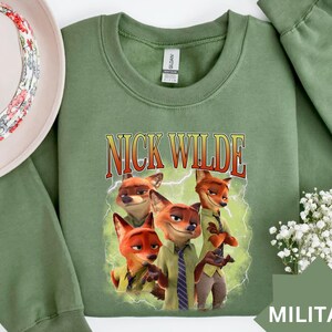 May include: A military green sweatshirt featuring a graphic of Nick Wilde from Zootopia. The design includes the text "NICK WILDE" in a vintage font, with illustrations of the character. The sweatshirt is a crew neck style.