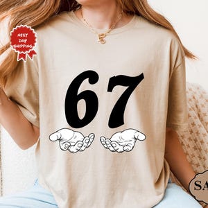 May include: Sand-colored t-shirt featuring the number "67" in bold black font above two open hands. The shirt has a round neckline and short sleeves. The image also includes a "Next Day Shipping" tag.