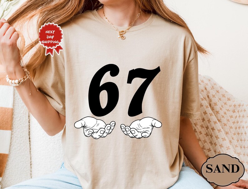 May include: Sand-colored t-shirt featuring the number "67" in bold black font above two open hands. The shirt has a round neckline and short sleeves. The image also includes a "Next Day Shipping" tag.
