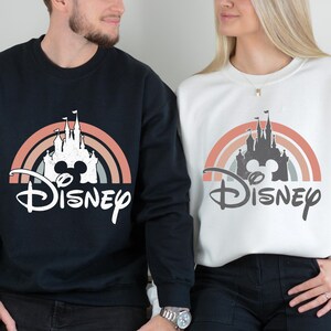 Puede incluir: Una sudadera negra y una sudadera blanca, cada una con un estampado gr&aacute;fico de la silueta de un castillo centrada dentro de un arco&iacute;ris semicircular multicolor, con la palabra Disney escrita en fuente cursiva debajo del dise&ntilde;o del castillo.