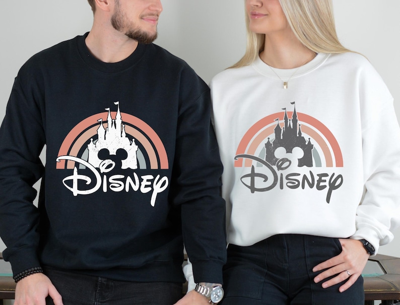 Puede incluir: Una sudadera negra y una sudadera blanca, cada una con un estampado gr&aacute;fico de la silueta de un castillo centrada dentro de un arco&iacute;ris semicircular multicolor, con la palabra Disney escrita en fuente cursiva debajo del dise&ntilde;o del castillo.
