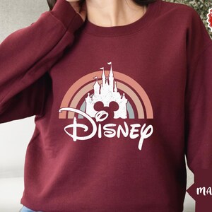 Puede incluir: Una sudadera de manga larga color granate con un gr&aacute;fico blanco de la silueta de un castillo centrado dentro de un arco&iacute;ris de tonos apagados. Debajo del castillo, la palabra Disney est&aacute; impresa en una fuente cursiva blanca con aspecto desgastado.