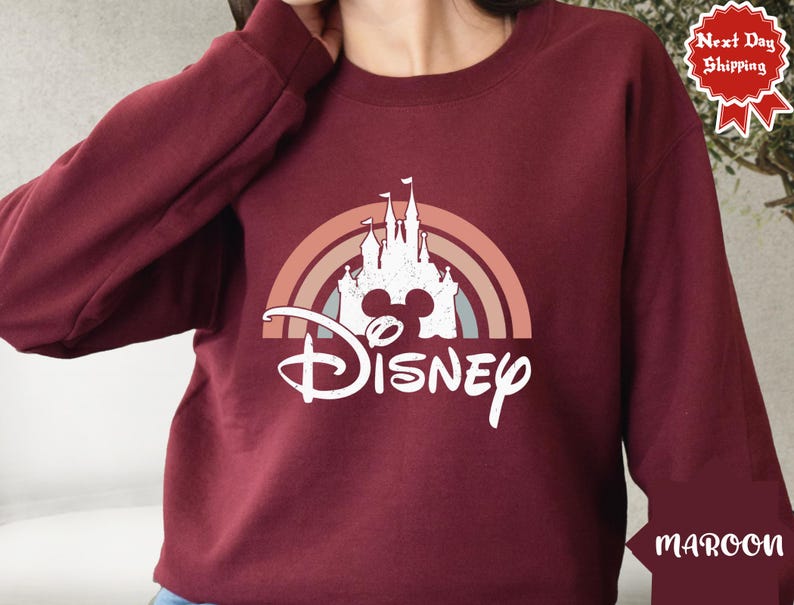 Puede incluir: Una sudadera de manga larga color granate con un gr&aacute;fico blanco de la silueta de un castillo centrado dentro de un arco&iacute;ris de tonos apagados. Debajo del castillo, la palabra Disney est&aacute; impresa en una fuente cursiva blanca con aspecto desgastado.