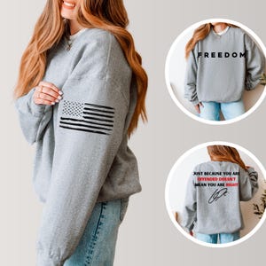 Op de afbeelding: Een grijze sweatshirt met een verweerde Amerikaanse vlag op de linkermouw. Extra afbeeldingen tonen het woord "FREEDOM" op de voorkant en een citaat op de achterkant: "JUST BECAUSE YOU ARE OFFENDED DOESN'T MEAN YOU ARE RIGHT."