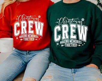 May include: Two sweatshirts, one red and one green, with the text "Christmas CREW MAKING MEMORIES TOGETHER" in white. The red sweatshirt is held by a person holding a white mug. Both people are wearing blue jeans.