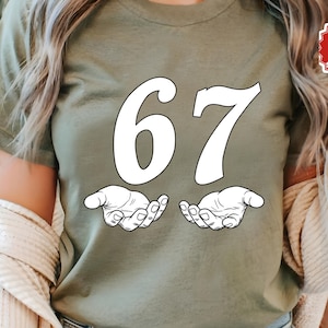 May include: Olive green t-shirt featuring the number "67" in white, with a stylized hand graphic beneath each number. The shirt has a crew neck and short sleeves. The image also includes a "Next Day Shipping" sticker.