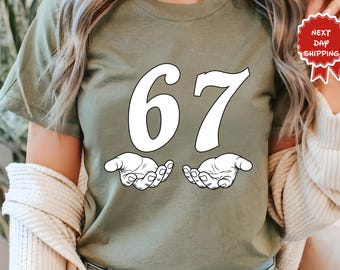 May include: Olive green t-shirt featuring the number "67" in white, with a stylized hand graphic beneath each number. The shirt has a crew neck and short sleeves. The image also includes a "Next Day Shipping" sticker.