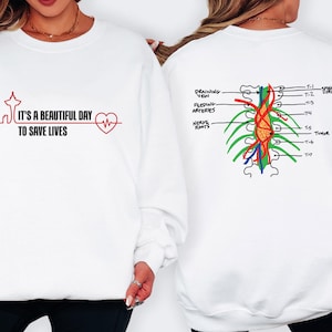 Op de afbeelding: Witte sweatshirt met de tekst "It's a beautiful day to save lives" in zwart en rood, samen met een hartslag grafiek en een skyline van Seattle. De achterkant toont een medische illustratie van de wervelkolom.