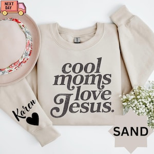 May include: A sand-colored sweatshirt with the text "cool moms love Jesus" in a dark gray font. The sleeve has the name "Karen" and a black heart. A hat with a floral band is on the left. A bouquet of white flowers is on the right.