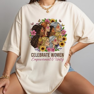 May include: Cream-colored t-shirt featuring a graphic of diverse women surrounded by flowers and butterflies. The text "CELEBRATE WOMEN Empowerment & Unity" is printed below the graphic. The shirt has short sleeves and a relaxed fit.