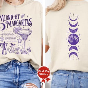 Puede incluir: Una camiseta beige con un estampado gráfico morado. La parte delantera muestra el texto Midnight Margaritas, una copa de cóctel, una rana con sombrero de bruja e ilustraciones botánicas. La parte trasera muestra fases lunares.