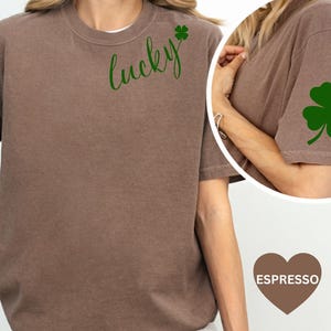 May include: Espresso-colored t-shirt with the word "lucky" in green script and a green shamrock on the sleeve. The shirt has a crew neck and short sleeves. The shirt is a casual style.