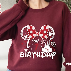 May include: Maroon sweatshirt with a white graphic of Minnie Mouse ears, a bow with red polka dots, a castle, fireworks, and the text "My 7th Birthday".