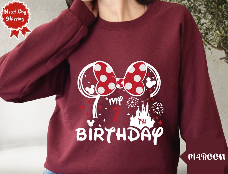 May include: Maroon sweatshirt with a white graphic of Minnie Mouse ears, a bow with red polka dots, a castle, fireworks, and the text "My 7th Birthday".