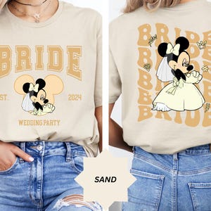 Può includere: Una maglietta color sabbia con un design da sposa con una Minnie Mouse dei cartoni animati che indossa un abito da sposa bianco e un velo. Il testo sulla maglietta recita "BRIDE EST. 2024 WEDDING PARTY".