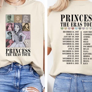 May include: A beige t-shirt with a graphic of Disney princesses and the text "Princess The Eras Tour". The graphic includes Snow White, Cinderella, Aurora, Ariel, Belle, Jasmine, Pocahontas, Mulan, Tiana, Rapunzel, Merida, Elsa and Anna, Moana, and Raya. The dates of their respective films are listed on the right side of the shirt.