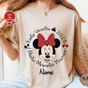 May include: A sand-coloured t-shirt with a Minnie Mouse graphic, sporting a red polka-dot bow. The design includes the text "Family Vacation 2025 Making Memories Together" and space for a name. A "Next Day Shipping" sticker is visible.