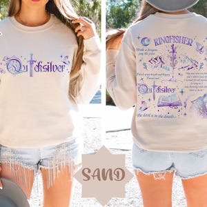 K&ouml;nnte beinhalten: Sandfarbenes Sweatshirt mit dem Wort "Quicksilver" in einer stilisierten Schrift auf der Vorderseite. Auf der R&uuml;ckseite befindet sich das Wort "Kingfisher" und ein Design mit Schwert, Bergen, Wolf und Text. Das Sweatshirt hat lange &Auml;rmel.