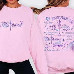 K&ouml;nnte beinhalten: Ein hellrosa Sweatshirt mit dem Wort "Quicksilver" und einer Schwertgrafik auf der Vorderseite. Auf der R&uuml;ckseite steht das Wort "Kingfisher" und verschiedene Illustrationen, darunter ein Gebirge, ein Wolf und Text.