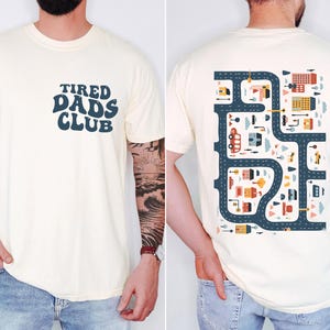 Può includere: T-shirt color crema con la scritta "TIRED DADS CLUB" in blu navy sul davanti. Il retro della maglietta presenta un'illustrazione colorata di una mappa stradale con edifici, auto e alberi.