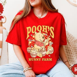 May include: A red t-shirt featuring a graphic of Winnie the Pooh with honey pots and the text "Pooh's Hunny Farm" in yellow. The shirt is a short-sleeved crew neck.