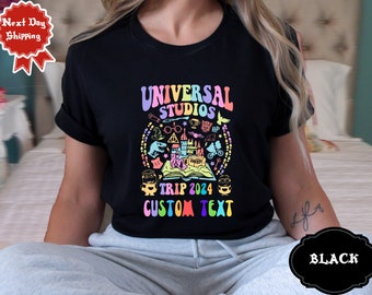May include: Black t-shirt featuring a colourful graphic of Universal Studios icons, including a castle, a book, and a minion. The text "Universal Studios Trip 2024 Custom Text" is printed below the graphic.
