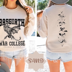 May include: A beige sweatshirt with a black dragon graphic and the text "Basgiath War College Dragon Riders" on the front. The back of the sweatshirt features a vertical line of five black dragons.