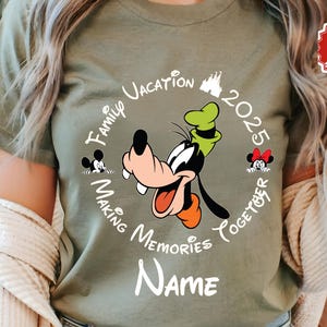 May include: Olive green t-shirt with a cartoon of Goofy and the text "Family Vacation 2025 Making Memories Together". The design includes Mickey and Minnie Mouse. The word "Name" is below the design.