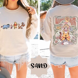 May include: A beige sweatshirt featuring a Winnie the Pooh graphic on the front and a larger Winnie the Pooh graphic on the back. The back graphic depicts Winnie the Pooh, Piglet, and Tigger within a floral design. The sweatshirt is modelled on a person wearing denim shorts.