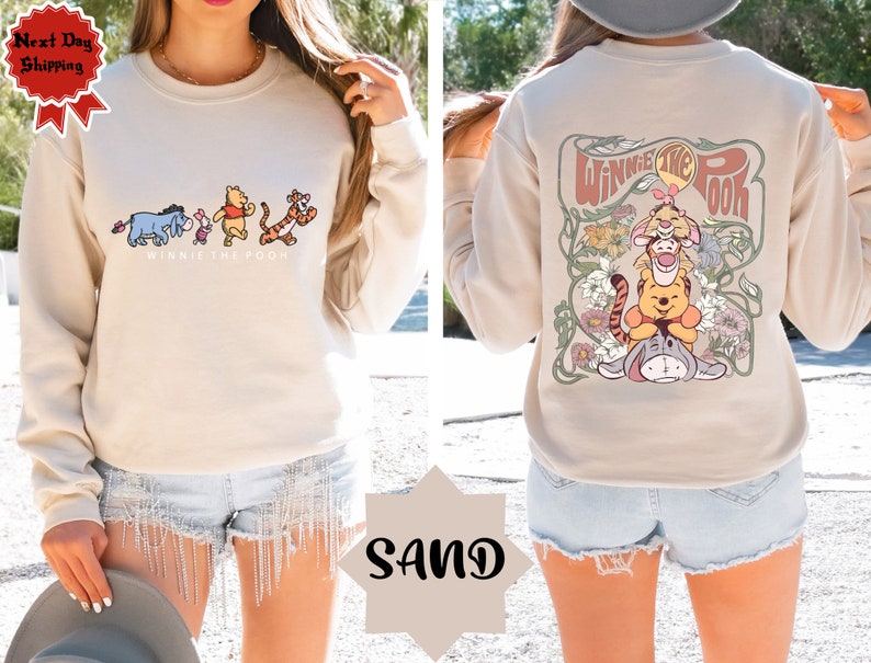 May include: A beige sweatshirt featuring a Winnie the Pooh graphic on the front and a larger Winnie the Pooh graphic on the back. The back graphic depicts Winnie the Pooh, Piglet, and Tigger within a floral design. The sweatshirt is modelled on a person wearing denim shorts.