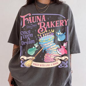 Może przedstawiać: Ciemnoszary t-shirt z nadrukiem graficznym zawierającym tekst Fauna Bakery Since 1959, Once Upon A Dream oraz Baked With Love & Magic. Wzór przedstawia wielowarstwowy tort ze świeczkami, miotłę oraz trzy małe postacie.