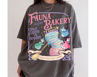 Può includere: Una t-shirt grigio antracite con una stampa grafica che riporta il testo Fauna Bakery Since 1959, Once Upon A Dream e Baked With Love & Magic. Il design include una torta a più strati con candeline, una scopa e tre piccole figure.