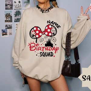 May include: A beige sweatshirt with a red and white Minnie Mouse bow, a black castle, and the text "NAME BIRTHDAY SQUAD 2021".