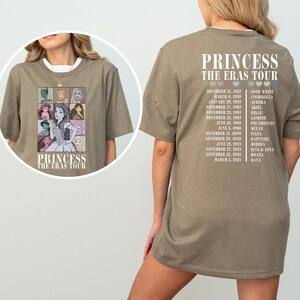 May include: A light brown t-shirt with a graphic of Disney princesses on the front and a list of tour dates and princess names on the back. The text on the shirt reads "Princess The Eras Tour".