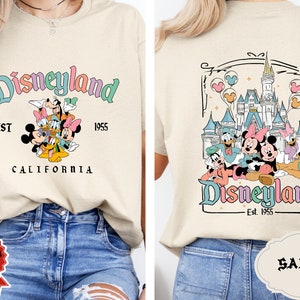 May include: A beige t-shirt with a colorful Disneyland graphic featuring Mickey Mouse, Minnie Mouse, Donald Duck, Goofy, and Pluto. The graphic includes the text "Disneyland Est. 1955 California". The back of the shirt features a colorful graphic of Sleeping Beauty's castle with Mickey Mouse, Minnie Mouse, Donald Duck, Goofy, and Pluto in front of it. The graphic includes the text "Disneyland Est. 1955".