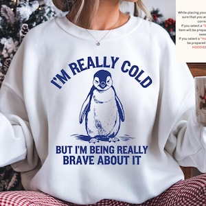 May include: White sweatshirt with a navy blue penguin graphic and the text "I'M REALLY COLD BUT I'M BEING REALLY BRAVE ABOUT IT." The sweatshirt also has a small text box with instructions. A mug with a holiday design is held in the hand.