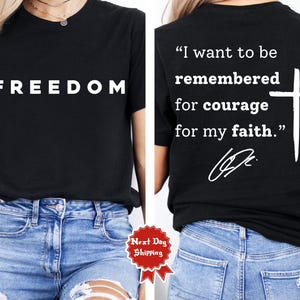 May include: Black t-shirt with the word "FREEDOM" in white block letters on the front. The back of the shirt features a white cross and the quote, "I want to be remembered for courage for my faith."