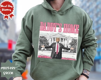 May include: A green hooded sweatshirt with a pink graphic print of a person standing in front of a building. The graphic text reads "Daddy's Home".
