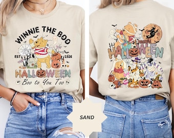 Op de afbeelding: Een beige T-shirt met een Winnie de Poeh Halloween-ontwerp. Het ontwerp toont Winnie de Poeh verkleed als een spook, met een pompoen in zijn hand en omringd door andere personages uit de Winnie de Poeh-franchise. De tekst "Winnie the Boo" is boven het ontwerp gedrukt, en de tekst "Pooh's Halloween" is onder het ontwerp gedrukt. De tekst "EST 1926" is aan de linkerkant van het ontwerp gedrukt, en de tekst "Boo to You Too" is onder het ontwerp gedruckt.
